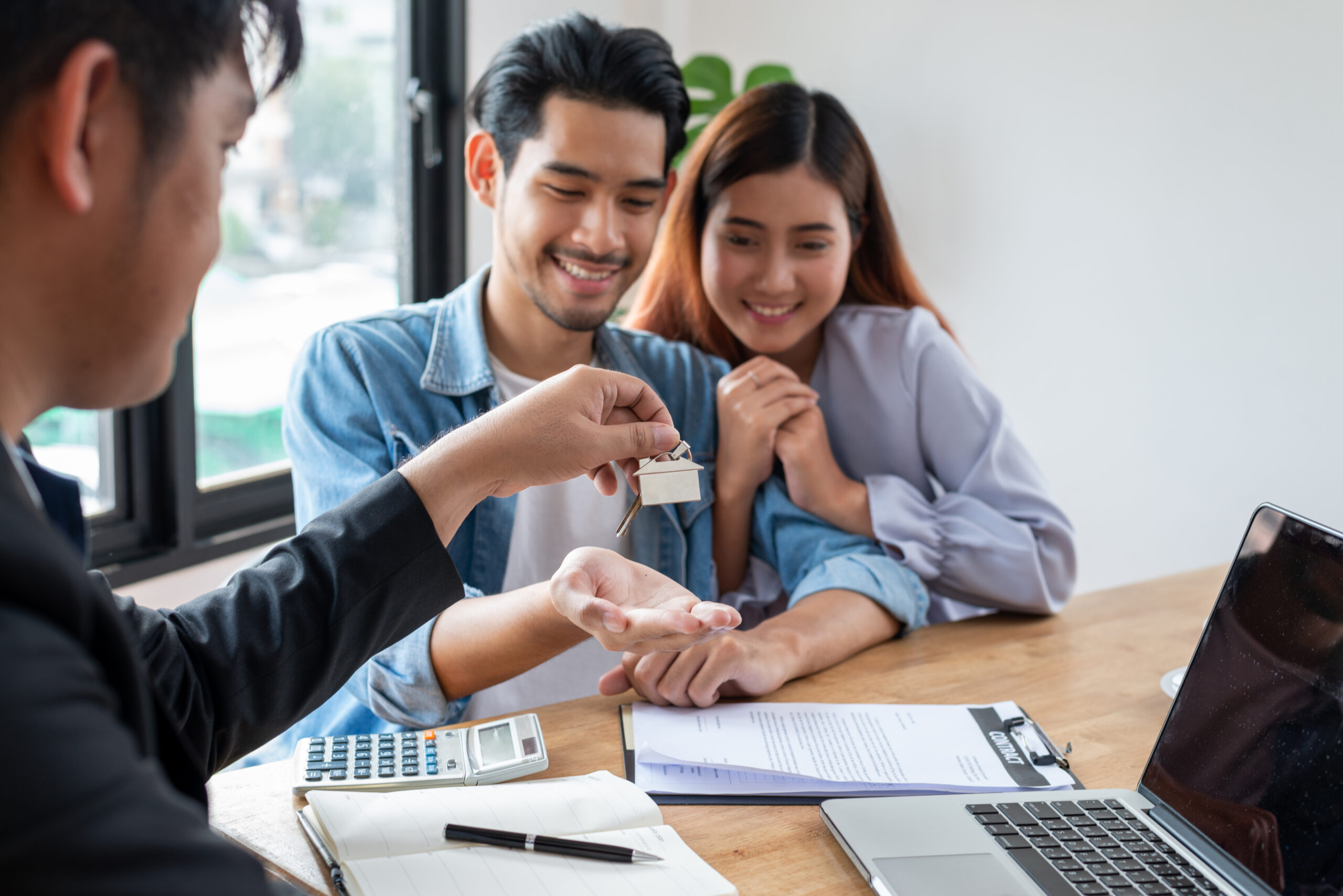 A couple excitedly receives house keys from a real estate agent during a home purchase meeting, with documents and a calculator on the table.