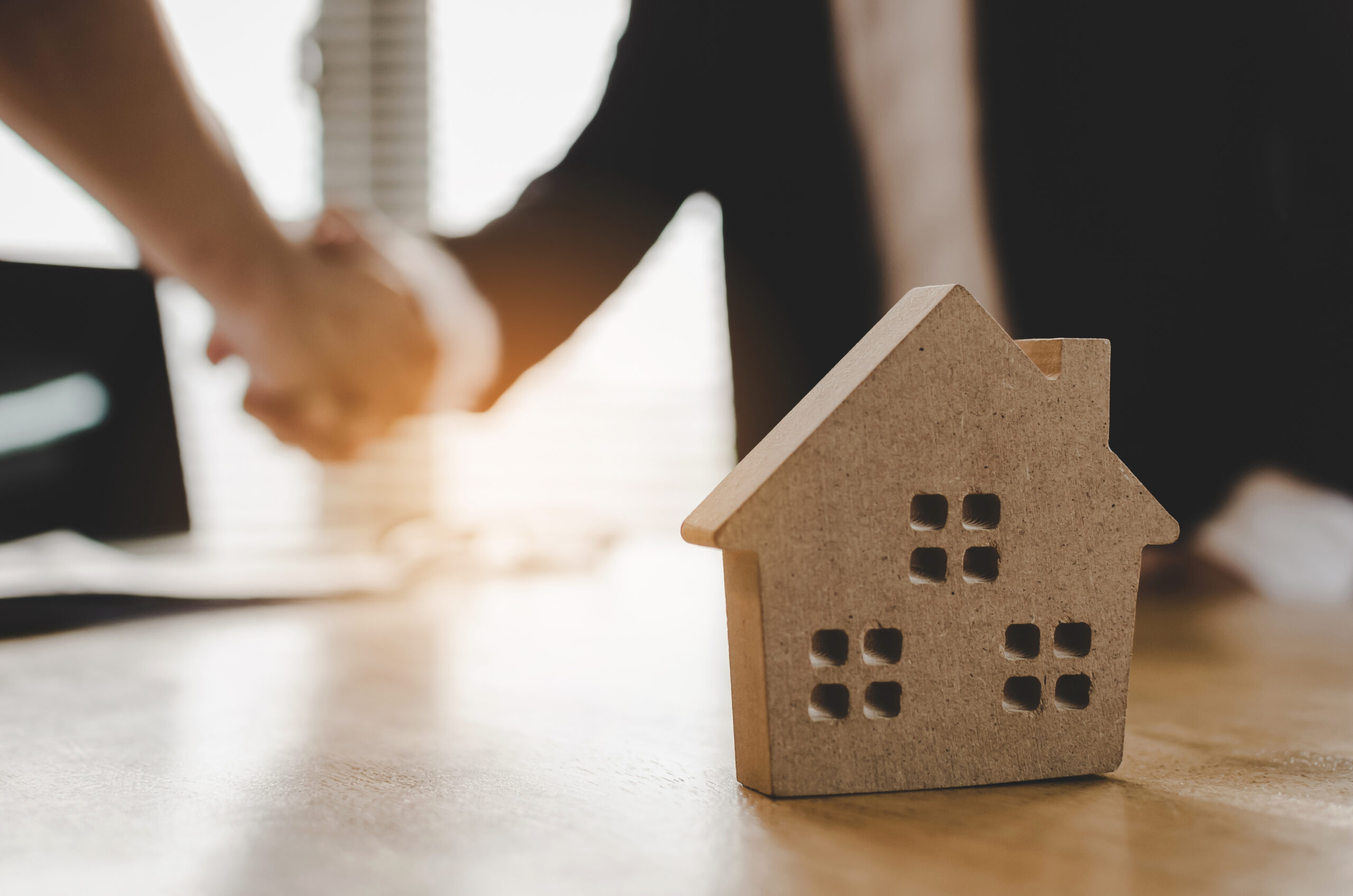 Two hands shake in a business setting, with a wooden house model in the foreground symbolizing a real estate transaction or agreement.