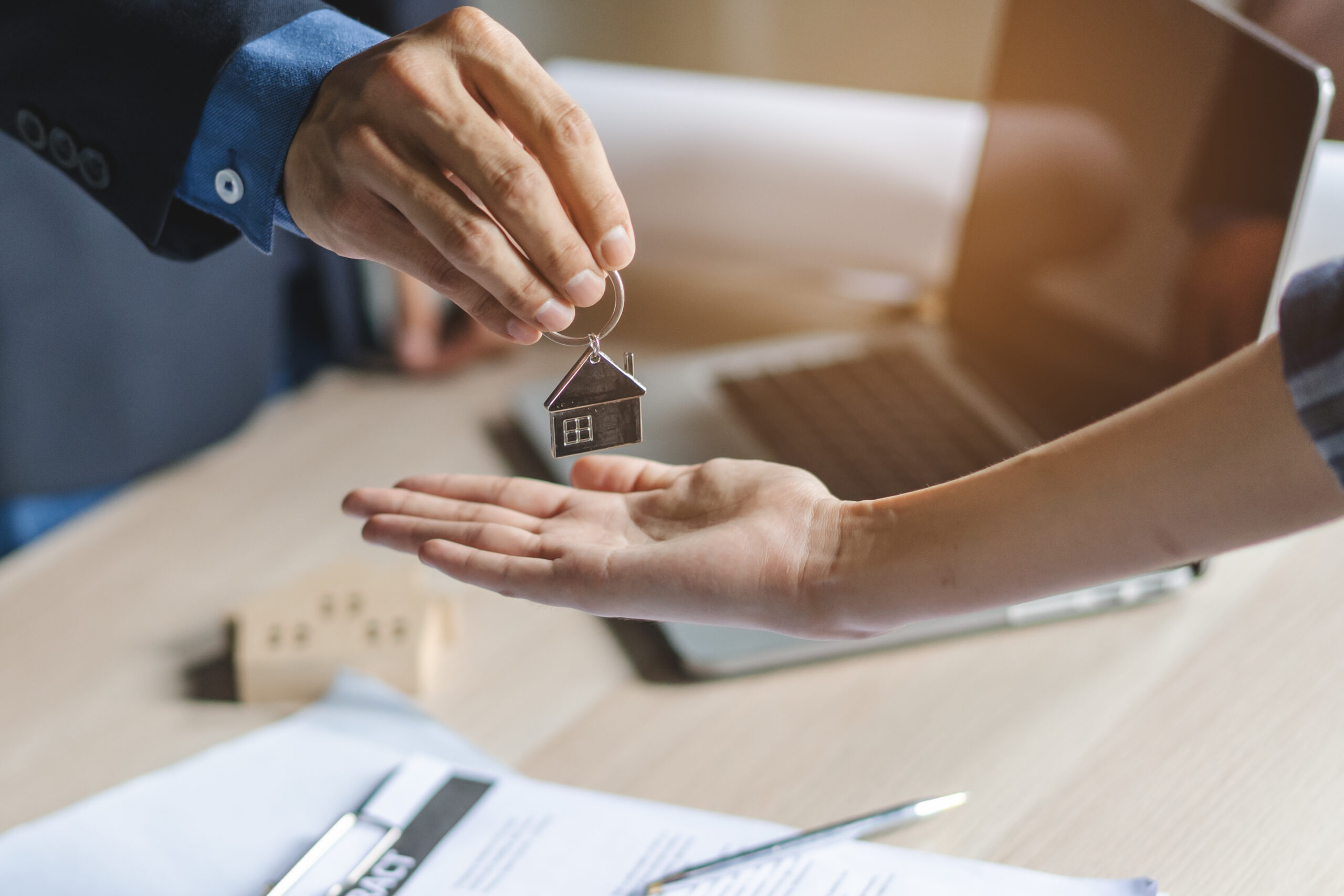 A key shaped like a house is being handed from one person to another, symbolizing a property transfer. Papers and a laptop are visible, indicating a real estate transaction.