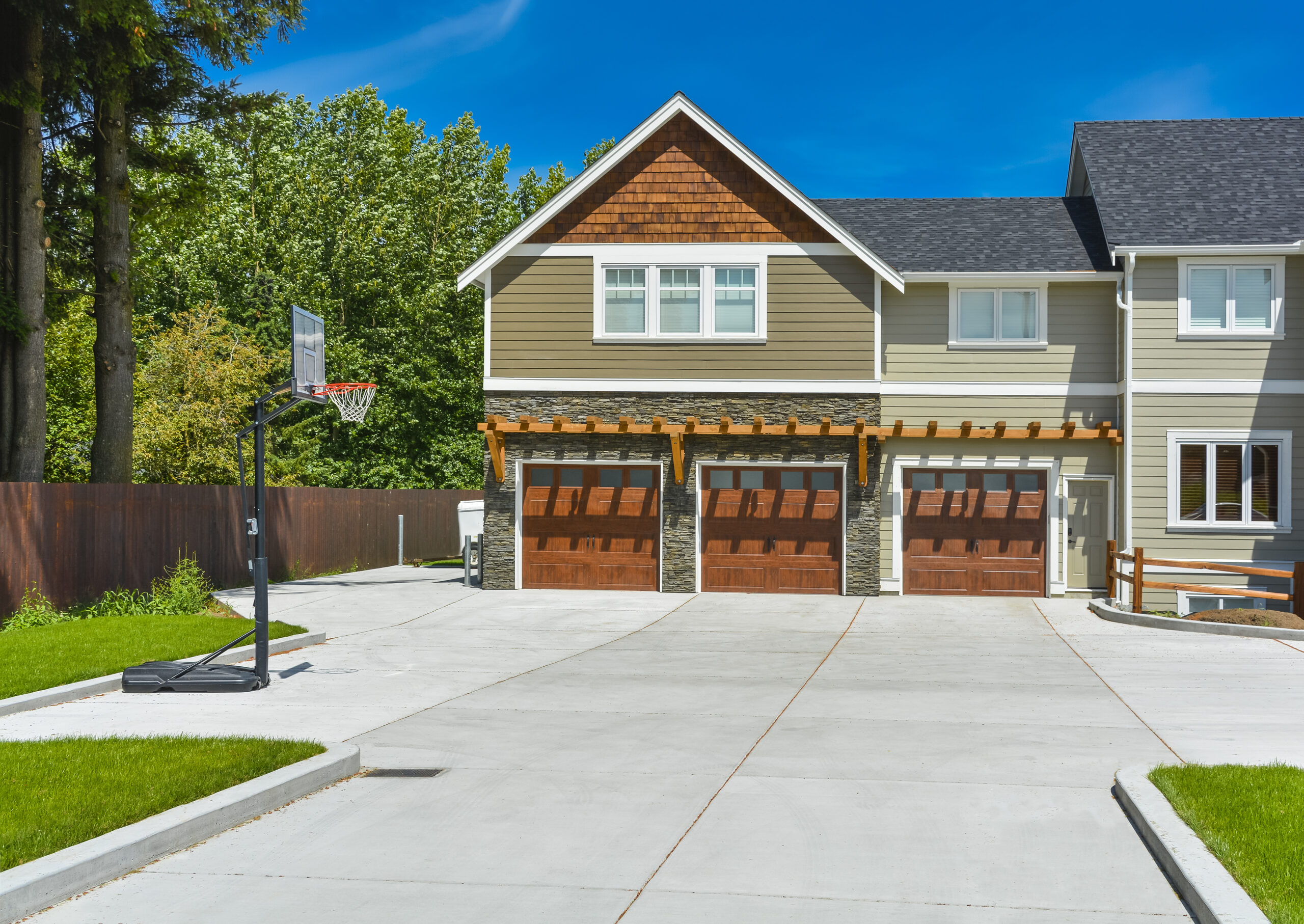 Modern home exterior featuring a garage with wooden doors and a basketball hoop on the driveway, surrounded by greenery and blue skies.