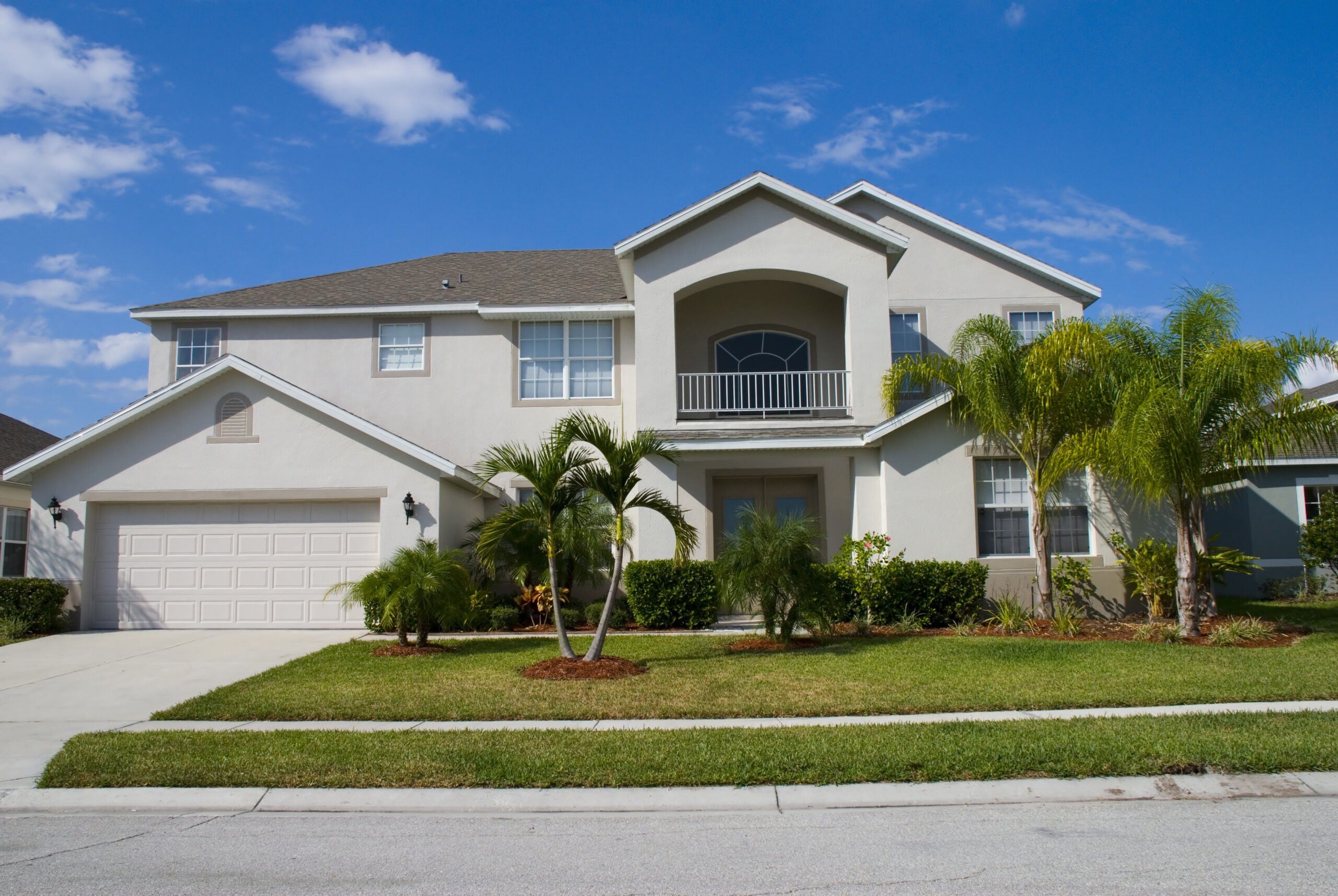 Two-story suburban home with a spacious front porch, palm trees, and a manicured lawn under a bright blue sky. Ideal for residential listings.