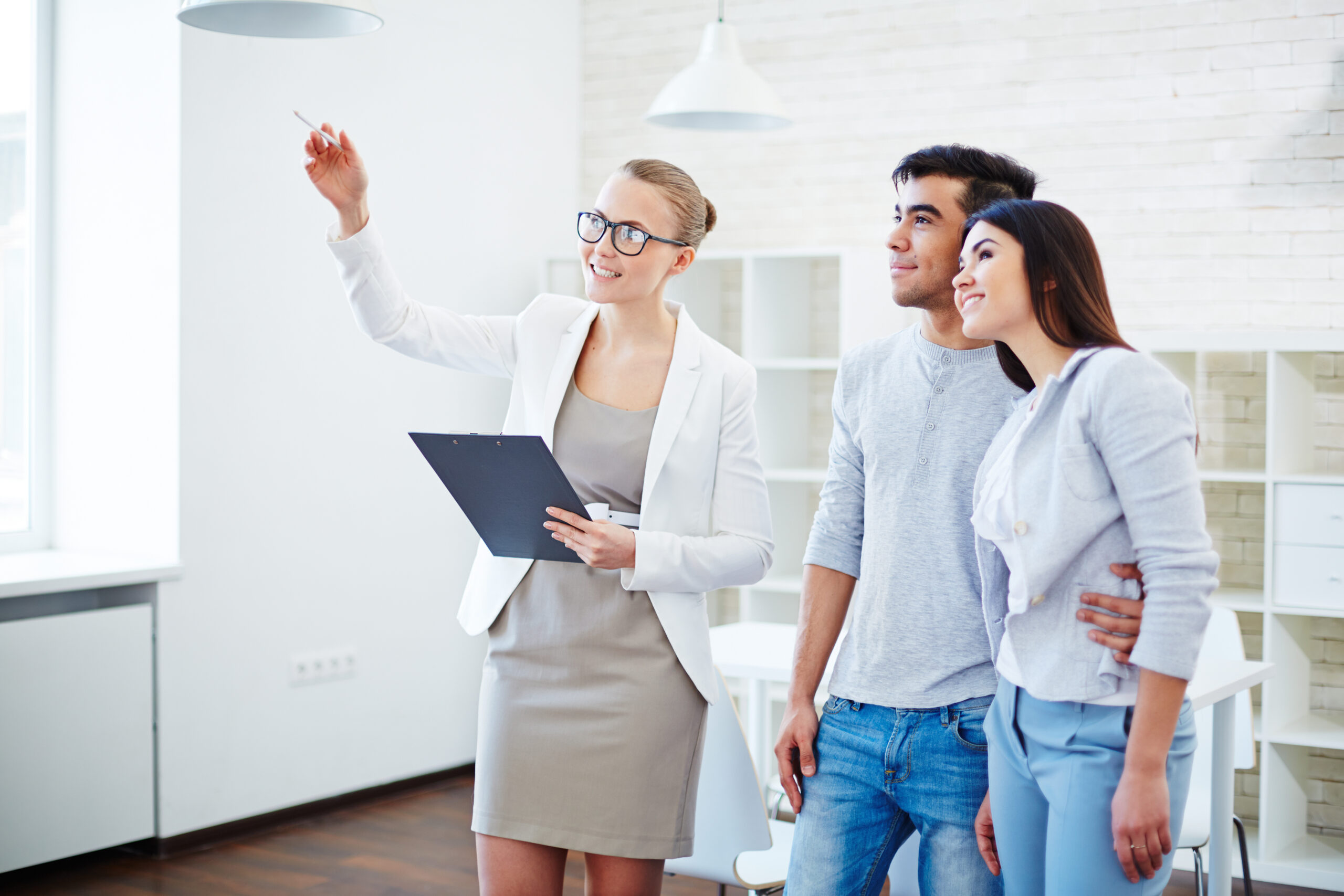 A professional woman gestures while explaining something to a couple, who are smiling and engaged in the conversation, in a modern office setting.