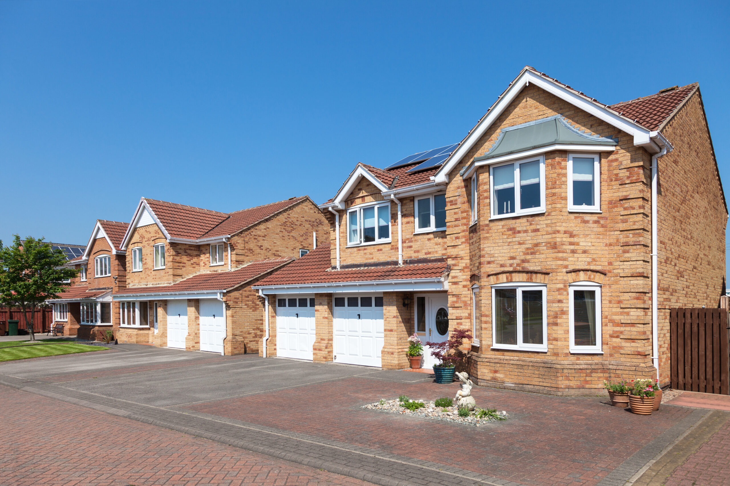 Modern brick houses lined along a residential street, featuring driveways, manicured lawns, and clear blue skies. Ideal for homebuyers.