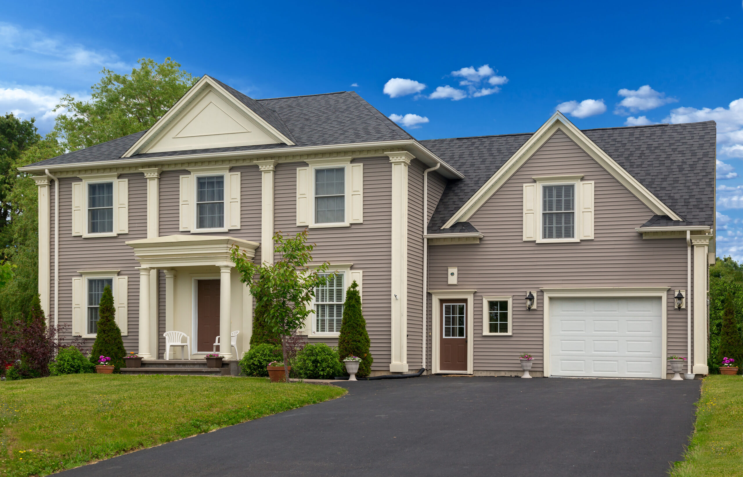 Two-story suburban home with light gray siding, white trim, and a front porch. Lush greenery and a driveway lead to a white garage door.